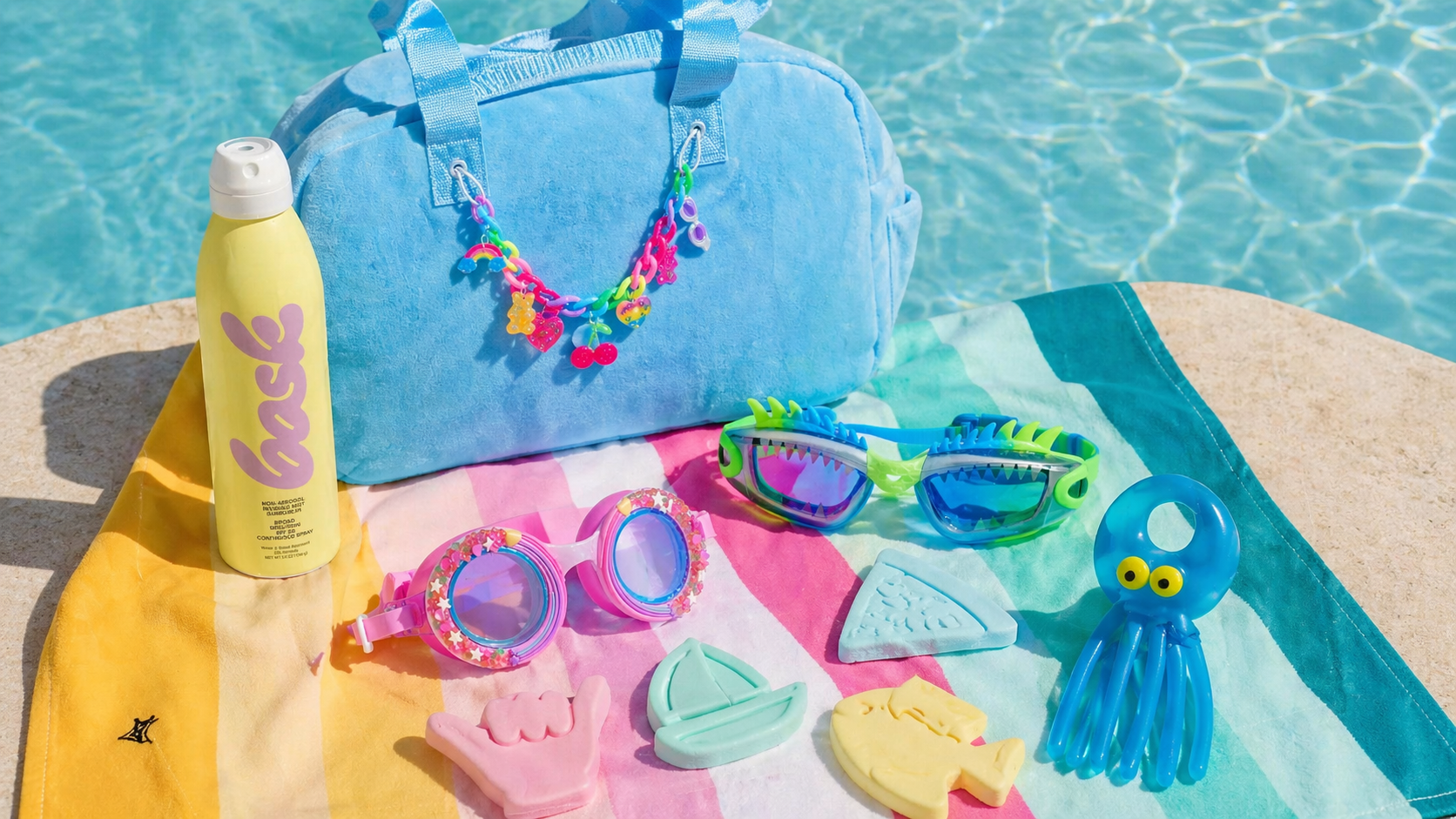 Poolside items including a blue bag, sunscreen bottle, goggles, and toys on a colorful towel by a pool.