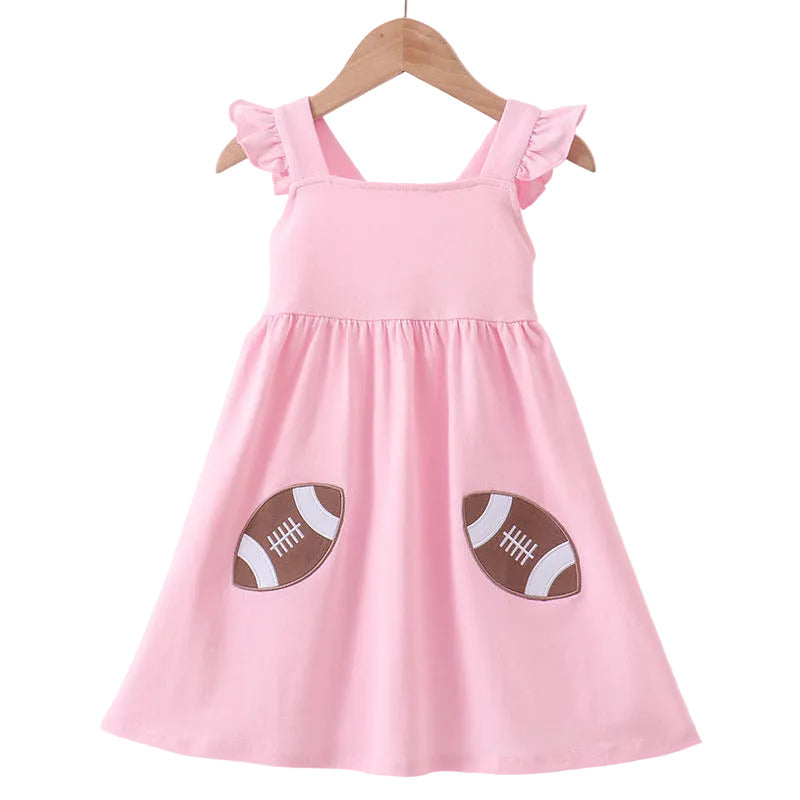 A pink sleeveless baby dress with ruffled shoulder straps and two football patches on the front, hanging on a wooden hanger against a light pink background.